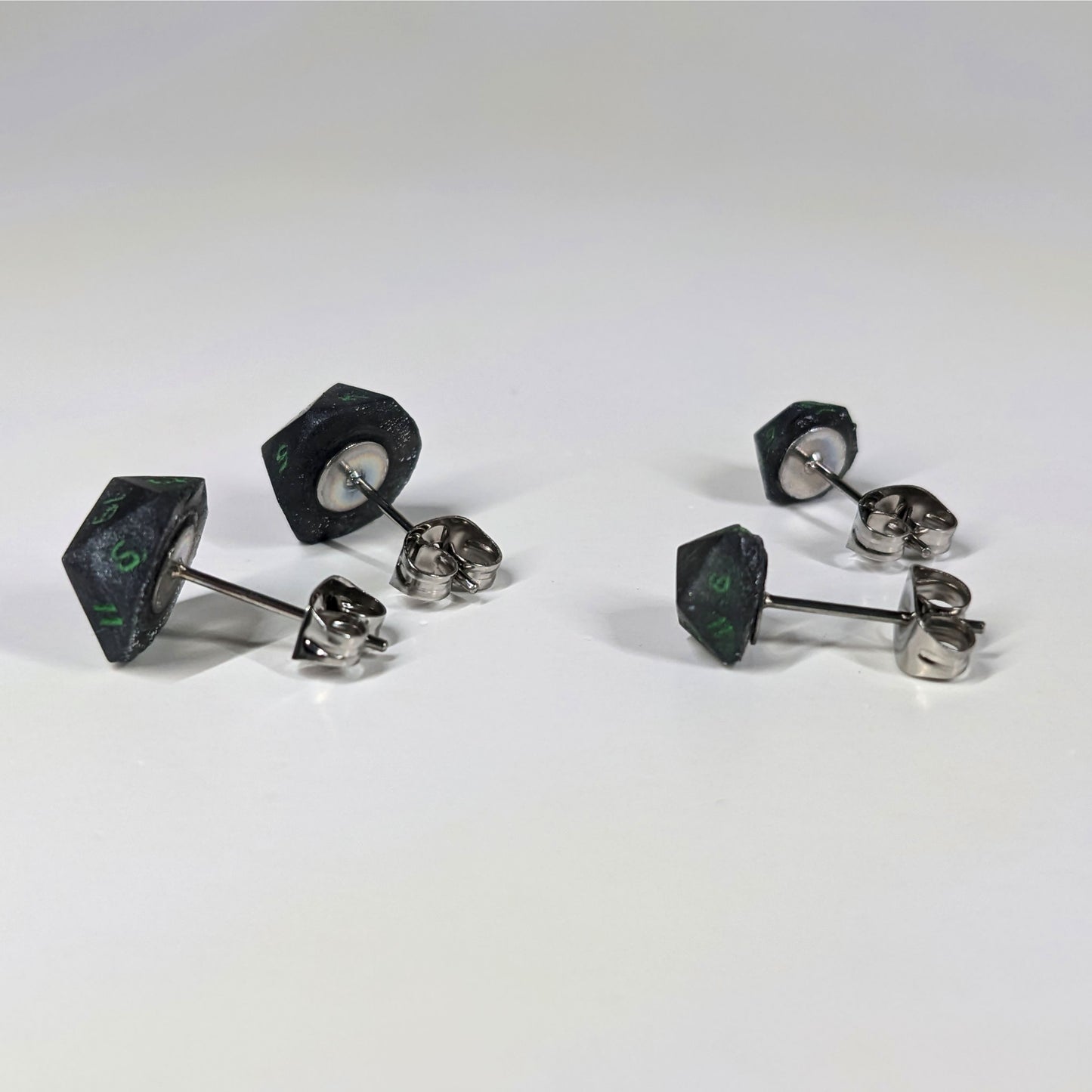 Two pairs of handmade d20 earrings that are black and silver with acid green numbers turned to show the metal alloy hardware and butterfly backs.