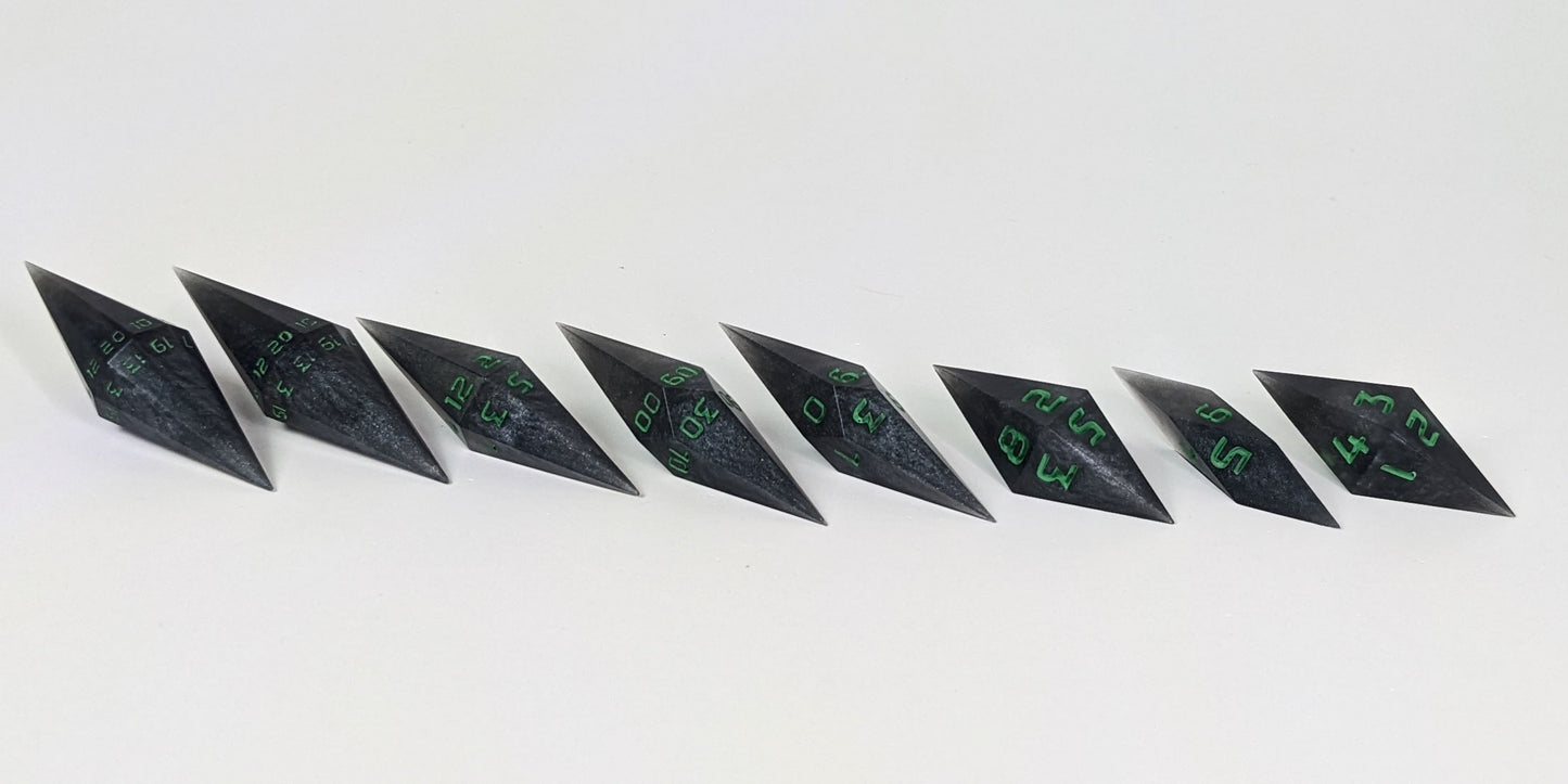 A mid angle photo of a complete set of handmade diamond DnD dice that are shadowy swirls of black and silver with acidic green numbers arranged in a straight line with bottom points angled slightly forwards and towards the right. They are lined up with the 2 d20s on the left going down to the d4 on the right side. The highest value number of each die is facing forward.