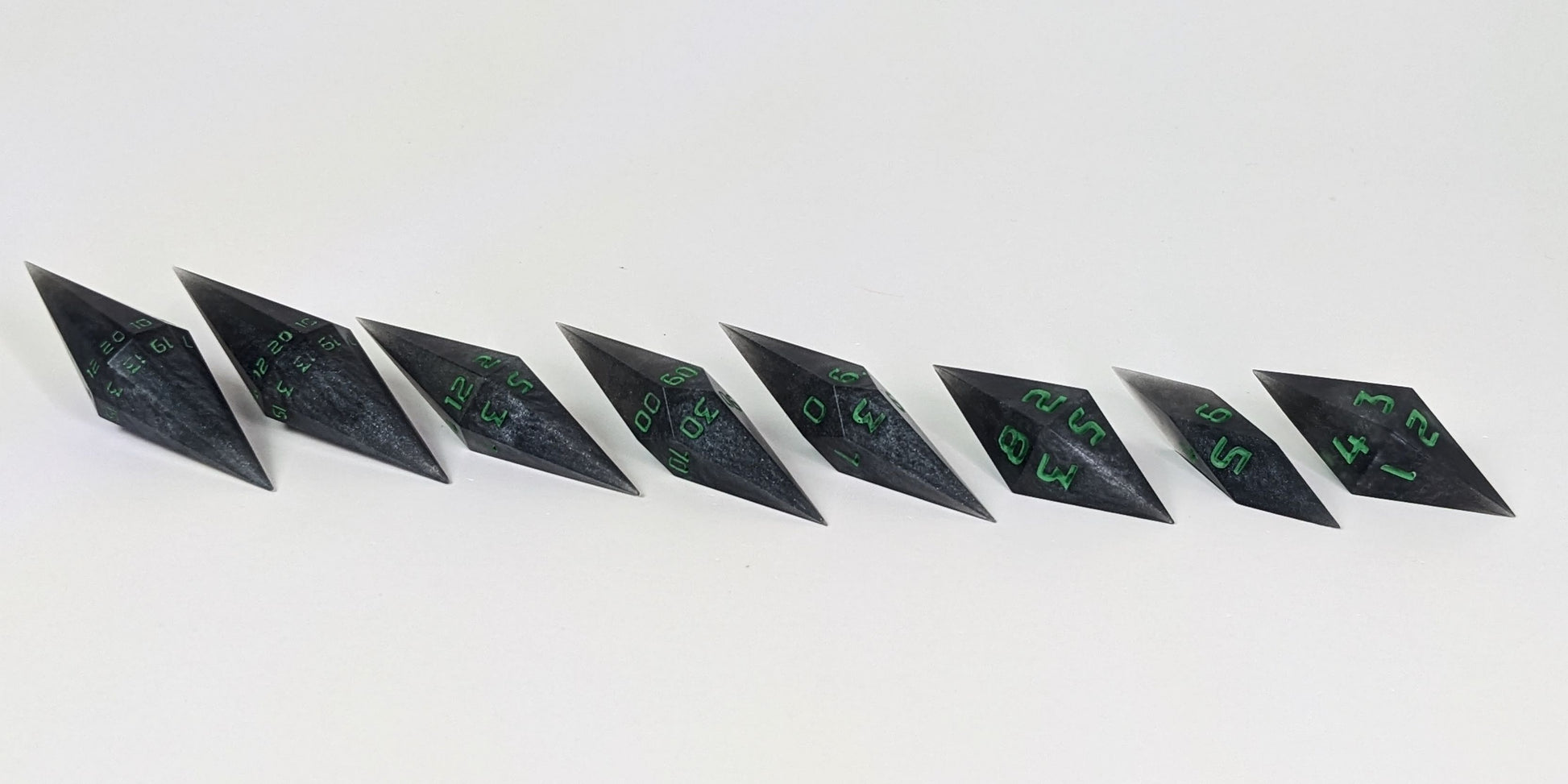 A mid angle photo of a complete set of handmade diamond DnD dice that are shadowy swirls of black and silver with acidic green numbers arranged in a straight line with bottom points angled slightly forwards and towards the right. They are lined up with the 2 d20s on the left going down to the d4 on the right side. The highest value number of each die is facing forward.