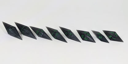 A mid angle photo of a complete set of handmade diamond DnD dice that are shadowy swirls of black and silver with acidic green numbers arranged in a straight line with bottom points angled slightly forwards and towards the right. They are lined up with the 2 d20s on the left going down to the d4 on the right side. The highest value number of each die is facing forward.