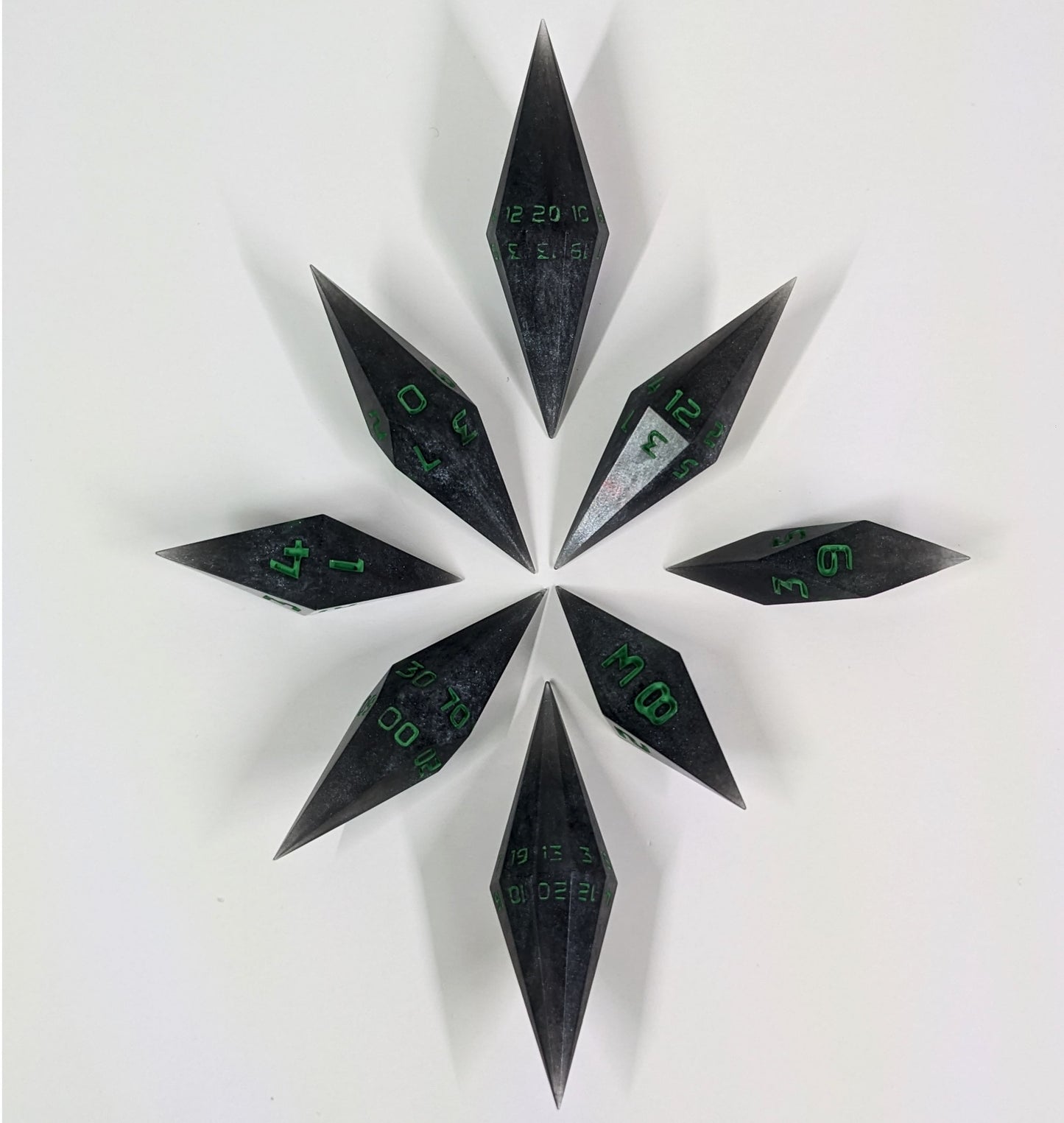 A complete set of diamond shaped resin dice featuring 2 d20s. These handmade dice shadowy swirls of black and silver with acidic green numbers and are clustered in a circle with the bottom points angled toward a central point on a white background.