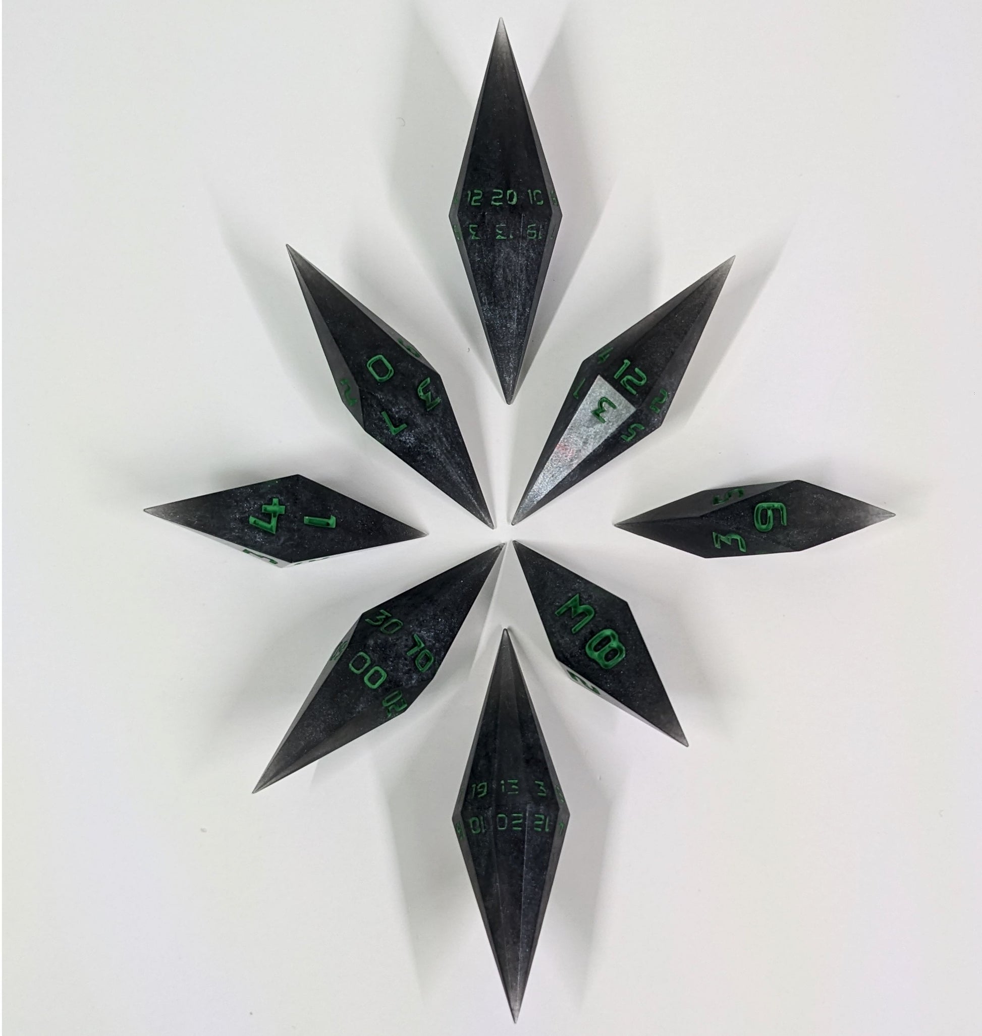 A complete set of diamond shaped resin dice featuring 2 d20s. These handmade dice shadowy swirls of black and silver with acidic green numbers and are clustered in a circle with the bottom points angled toward a central point on a white background.