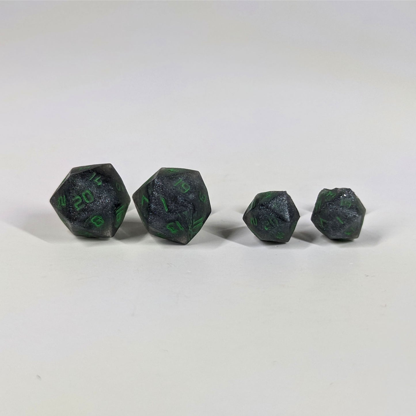 Two pairs of handmade d20 earrings with sterling silver hardware that are black and silver with acid green numbers on a white background sit in a line with the larger pair on the left and the smaller pair on the right. The earrings on the left of each pair centers the 20 face and the earrings on the right of each pair centers the 1 face.