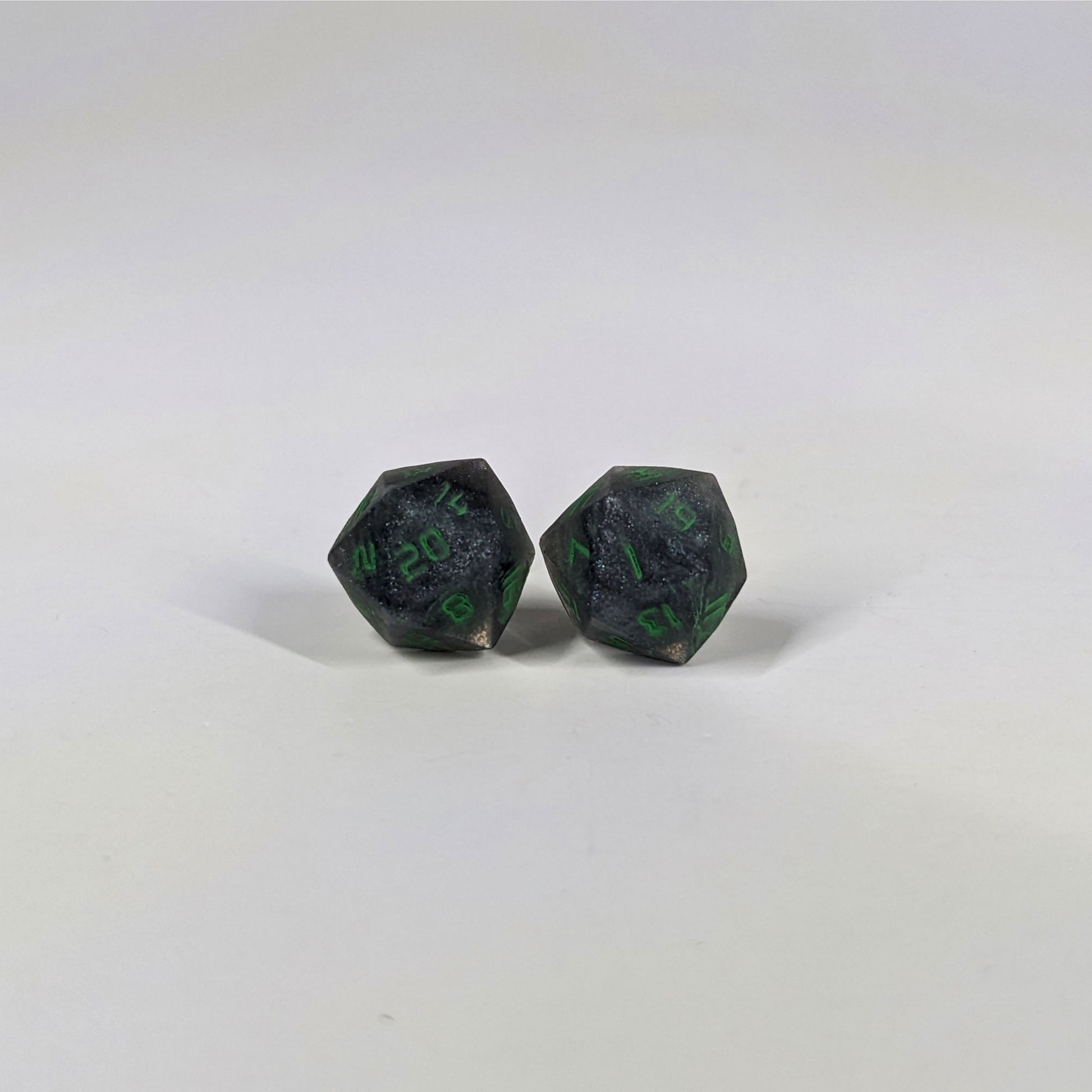 A single pair of the larger size handmade d20 earrings with sterling silver hardware that are black and silver with acid green numbers on a white background. The earring on the left centers the 20 face and the earring on the right centers the 1 face.