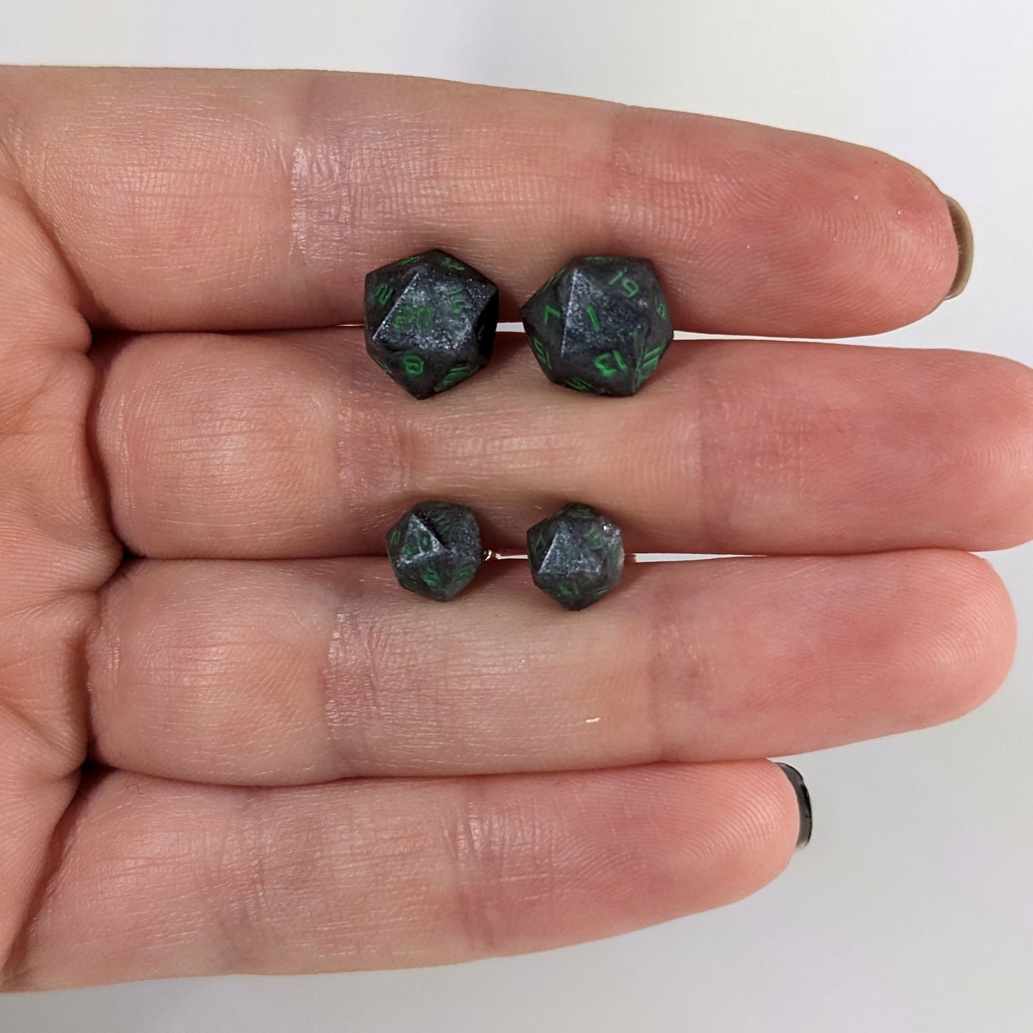 Two pairs of handmade d20 earrings with sterling silver hardware that are black and silver with acid green numbers held between the fingers of a flat hand. The larger pair is between the pointer and middle finger, the smaller between the middle and ring finger. The earrings on the left of each pair centers the 20 face and the earrings on the right of each pair centers the 1 face.