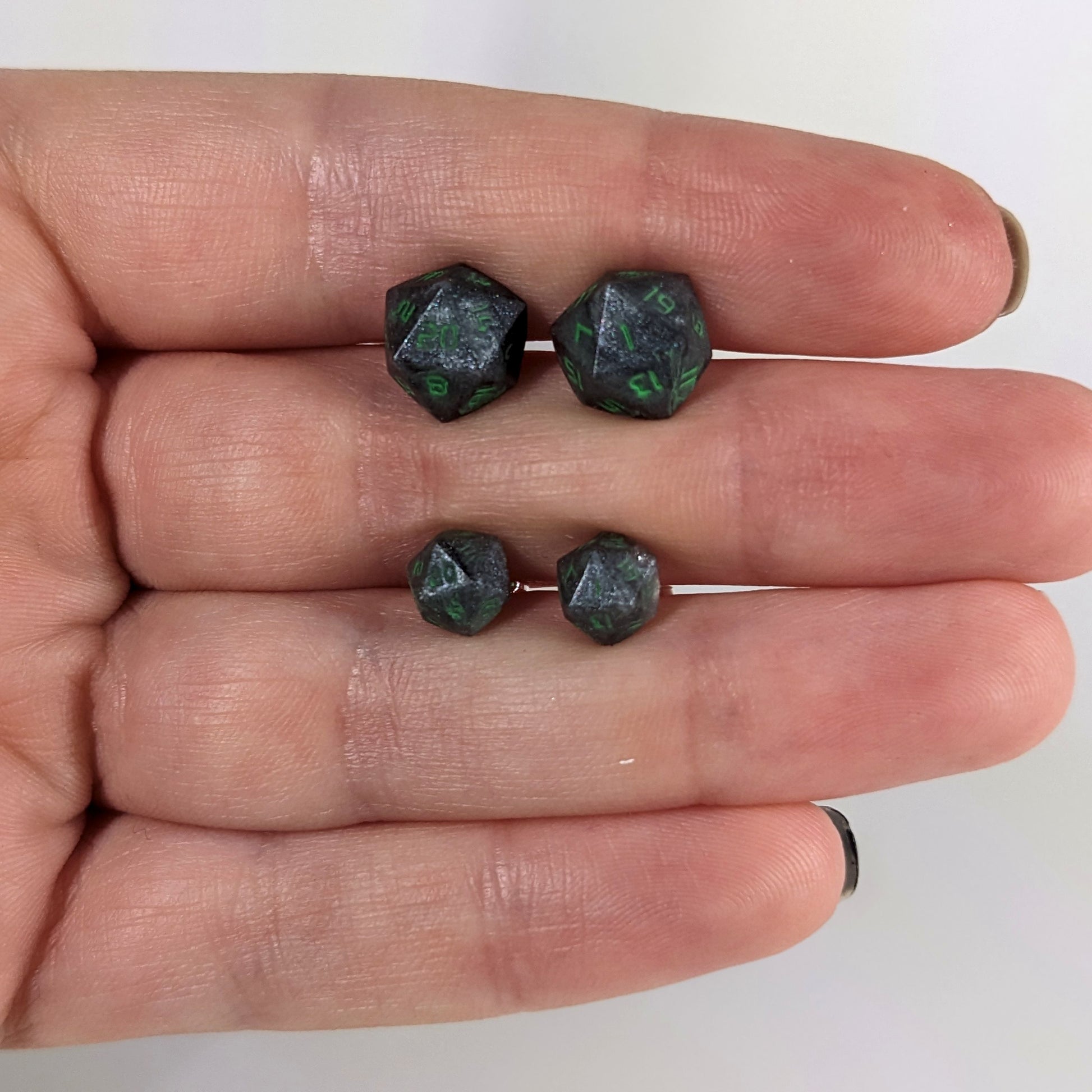 Two pairs of handmade d20 earrings with sterling silver hardware that are black and silver with acid green numbers held between the fingers of a flat hand. The larger pair is between the pointer and middle finger, the smaller between the middle and ring finger. The earrings on the left of each pair centers the 20 face and the earrings on the right of each pair centers the 1 face.