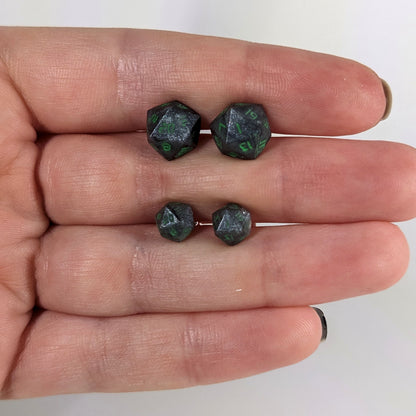 Two pairs of handmade d20 earrings with sterling silver hardware that are black and silver with acid green numbers held between the fingers of a flat hand. The larger pair is between the pointer and middle finger, the smaller between the middle and ring finger. The earrings on the left of each pair centers the 20 face and the earrings on the right of each pair centers the 1 face.
