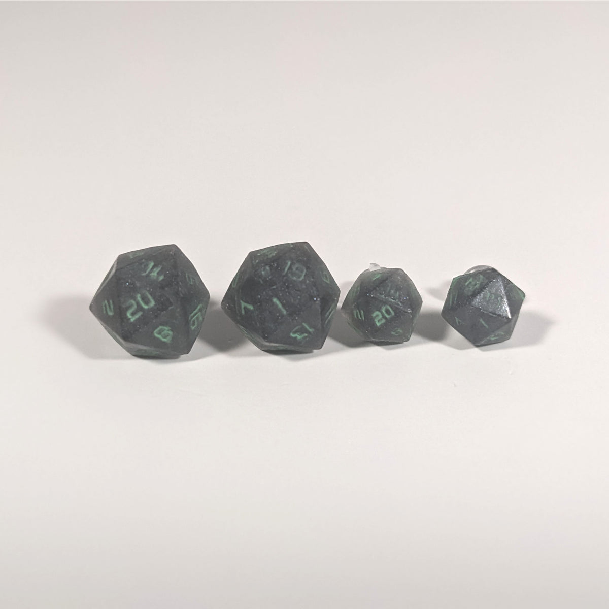 Two pairs of handmade d20 earrings with titanium hardware that are black with a touch of silver with acidic green numbers on a white background sit in a line with the larger pair on the left and the smaller pair on the right. The earrings on the left of each pair centers the 20 face and the earrings on the right of each pair centers the 1 face.