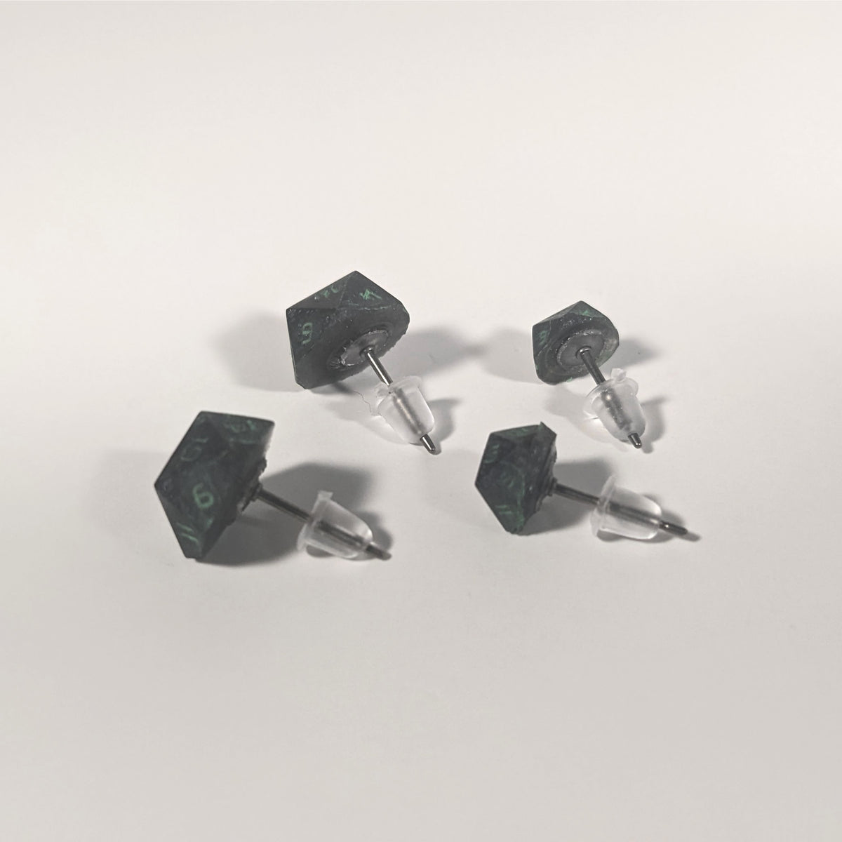 Two pairs of handmade d20 earrings that are black with a touch of silver with acidic green numbers turned to show the titanium hardware and bell shaped rubber backs.