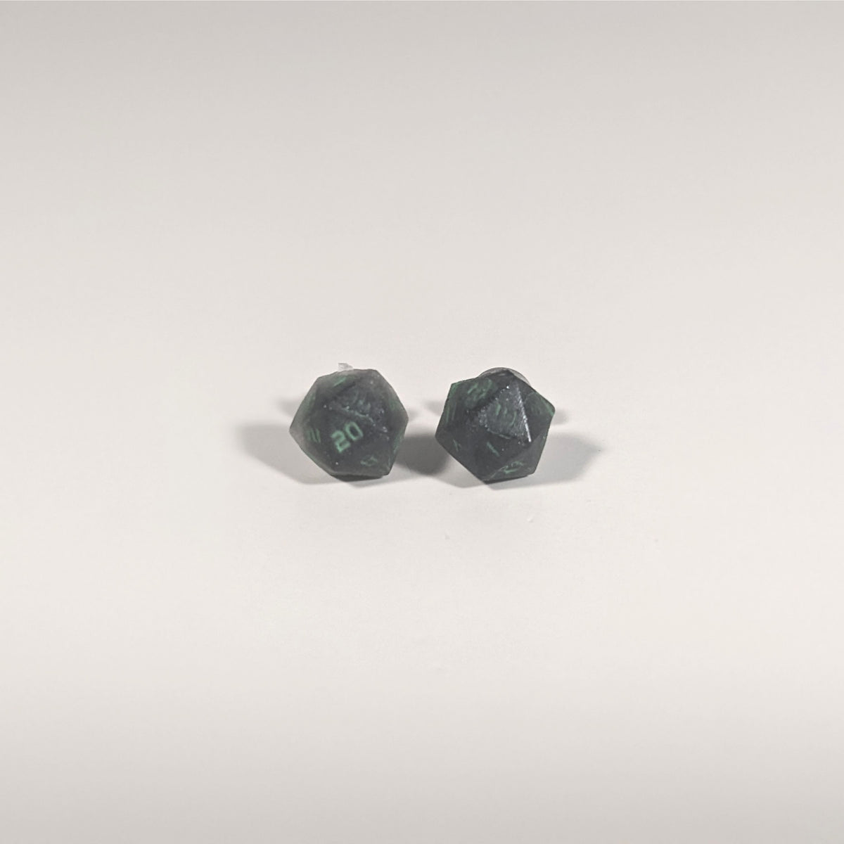 A single pair of the smaller size handmade d20 earrings with titanium hardware that are black with a touch of silver with acidic green numbers on a white background. The earring on the left centers the 20 face and the earring on the right centers the 1 face.