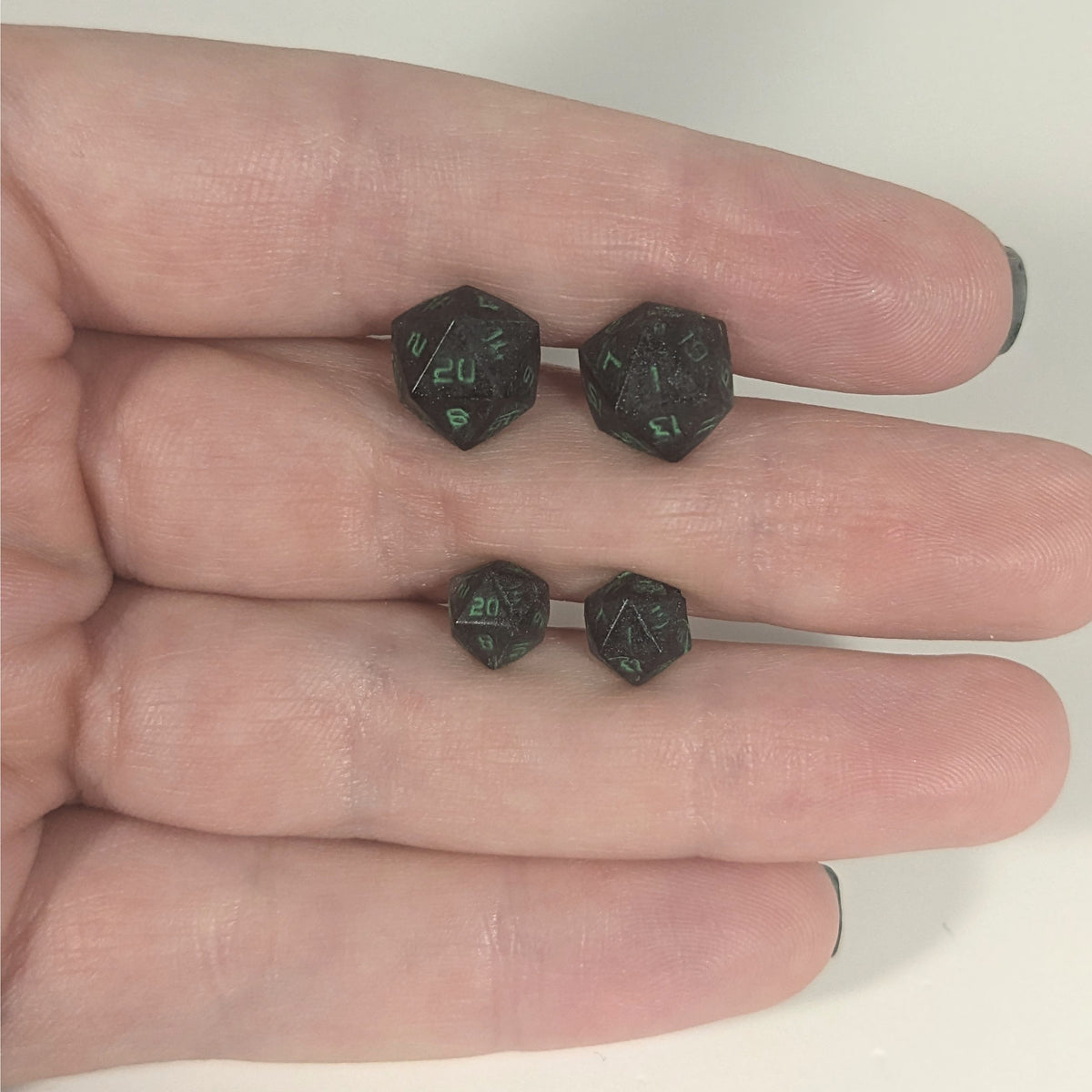 Two pairs of handmade d20 earrings with titanium hardware that are black with a touch of silver with acidic green numbers held between the fingers of a flat hand. The larger pair is between the pointer and middle finger, the smaller between the middle and ring finger. The earrings on the left of each pair centers the 20 face and the earrings on the right of each pair centers the 1 face.