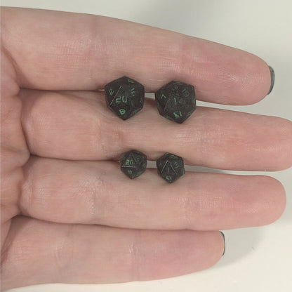 Two pairs of handmade d20 earrings with titanium hardware that are black with a touch of silver with acidic green numbers held between the fingers of a flat hand. The larger pair is between the pointer and middle finger, the smaller between the middle and ring finger. The earrings on the left of each pair centers the 20 face and the earrings on the right of each pair centers the 1 face.