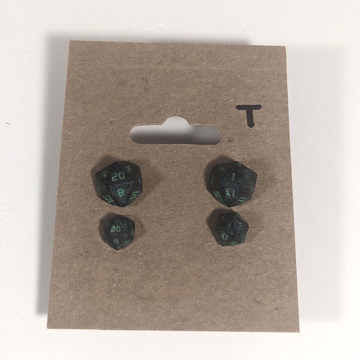 Two pairs of handmade d20 earrings with titanium hardware that are black with a touch of silver with acidic green numbers on a kraft brown earring card with a T in the top right corner. The earrings on the left of each pair centers the 20 face and the earrings on the right of each pair centers the 1 face.