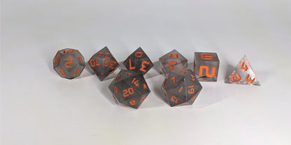 A straight on photo of a standard set of DnD dice that have black swirls and orange numbers focusing on 2 d20s in the center. The d20 on the left has the 20 face centered and the d20 on the right has the 1 face centered. The other 6 dice are lined up in the background.