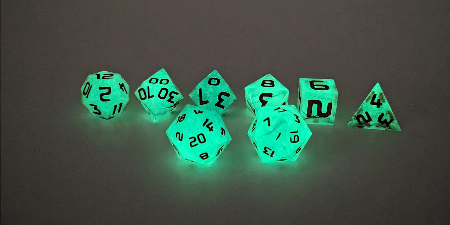A straight on photo of a standard set of DnD dice that have black swirls showing through glow-in-the-dark green and orange numbers focusing on 2 d20s in the center. The d20 on the left has the 20 face centered and the d20 on the right has the 1 face centered. The other 6 dice are lined up in the background.