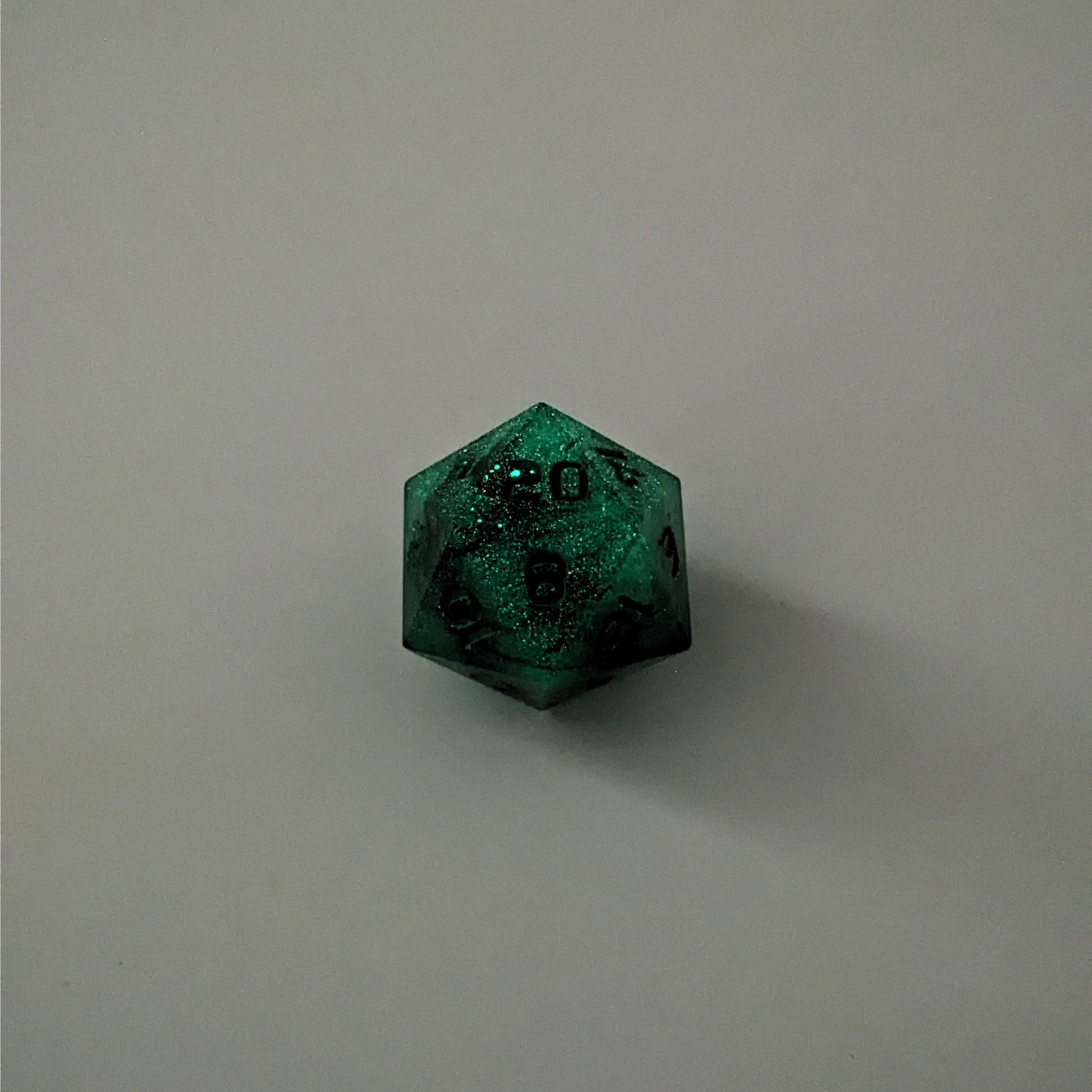 A high angle photo of a standard d20 glowing green with black swirls and green numbers with the 20 face up on a white background.