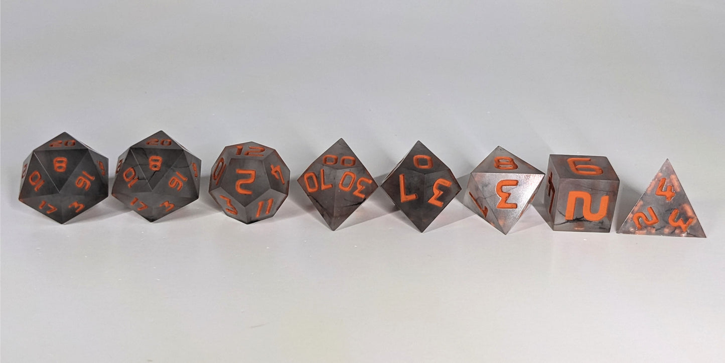 A straight on photo of a complete set of polyhedral dice that have black swirls and orange numbers arranged in a straight line. They are lined up with the 2 d20s on the left going down to the d4 on the right side. The highest value number of each die is facing up.