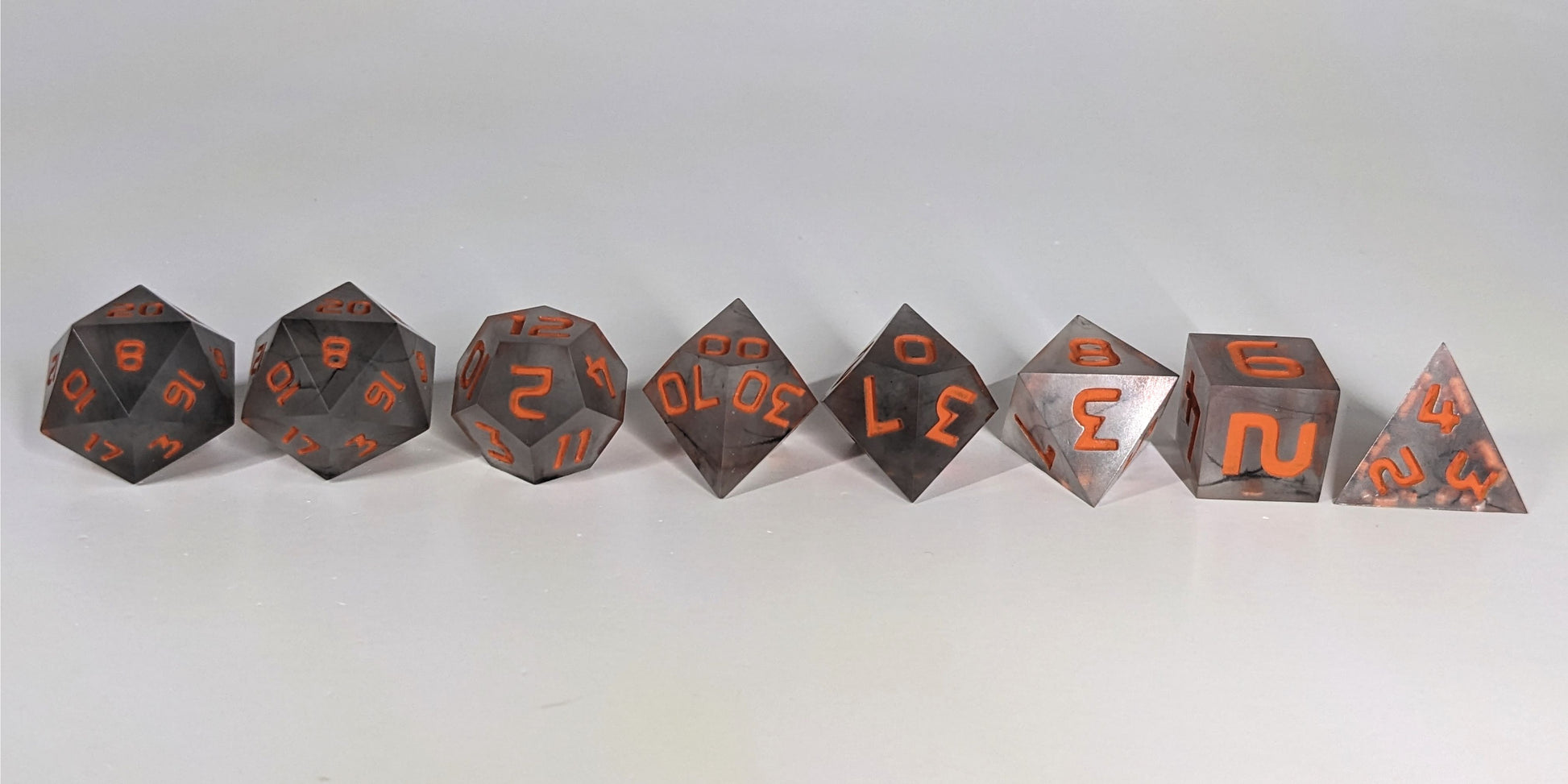 A straight on photo of a complete set of polyhedral dice that have black swirls and orange numbers arranged in a straight line. They are lined up with the 2 d20s on the left going down to the d4 on the right side. The highest value number of each die is facing up.