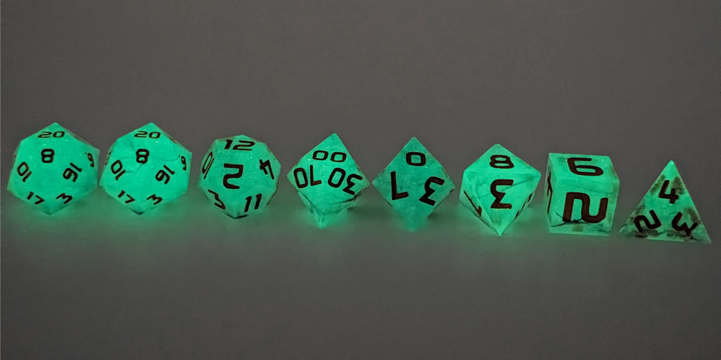 A straight on photo of a complete set of polyhedral dice that are glowing green and have orange numbers arranged in a straight line. They are lined up with the 2 d20s on the left going down to the d4 on the right side. The highest value number of each die is facing up. This image shows the glow-in-the-dark feature of this dice set and that you can still see the black swirls through the glow.