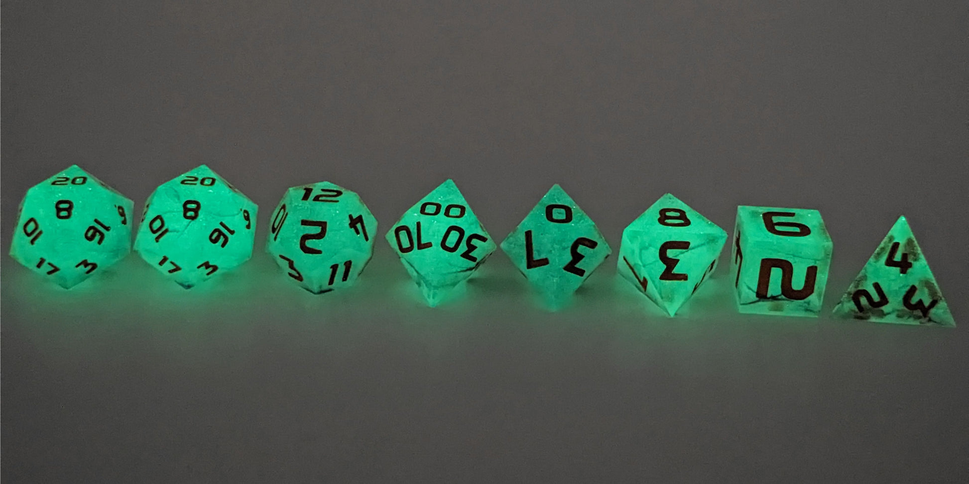 A straight on photo of a complete set of polyhedral dice that are glowing green and have orange numbers arranged in a straight line. They are lined up with the 2 d20s on the left going down to the d4 on the right side. The highest value number of each die is facing up. This image shows the glow-in-the-dark feature of this dice set and that you can still see the black swirls through the glow.
