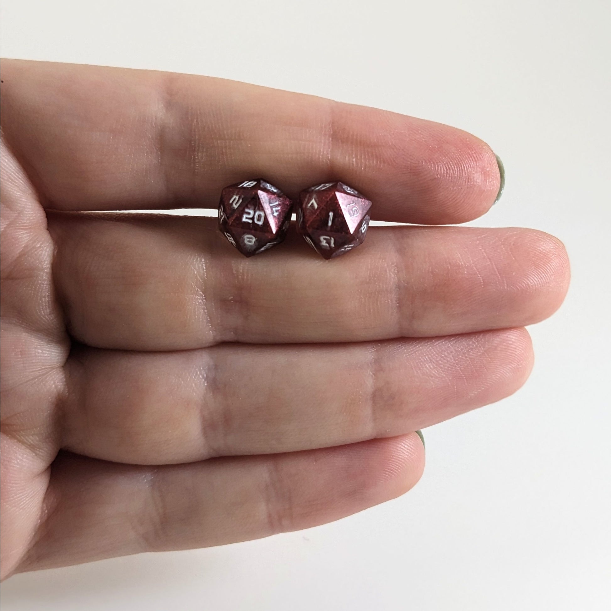A single pair of handmade d20 earrings that are swirls of red with white numbers held between the pointer and middle finger of a flat hand. The earring on the left centers the 20 face and the earring on the right centers the 1 face.