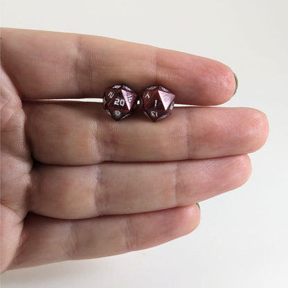 A single pair of handmade d20 earrings that are swirls of red with white numbers held between the pointer and middle finger of a flat hand. The earring on the left centers the 20 face and the earring on the right centers the 1 face.