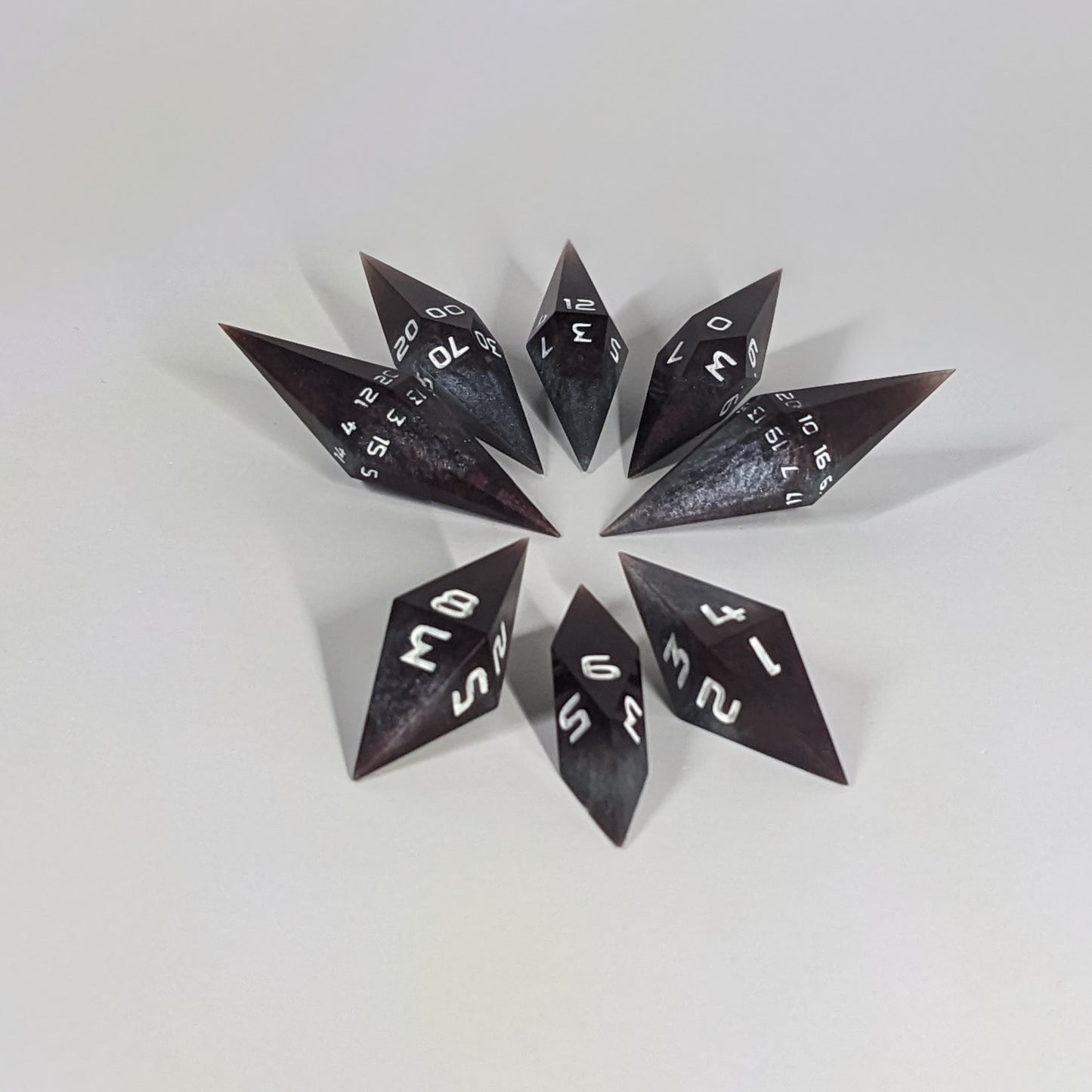 A complete set of resin dice featuring 2 d20s for advantage rolls. These deep maroon and dark silver with white numbers, slender handmade diamond dice are clustered in a tight circle on a white background, highlighting the quality of the artisan dice craftsmanship and the unique pointed shape.