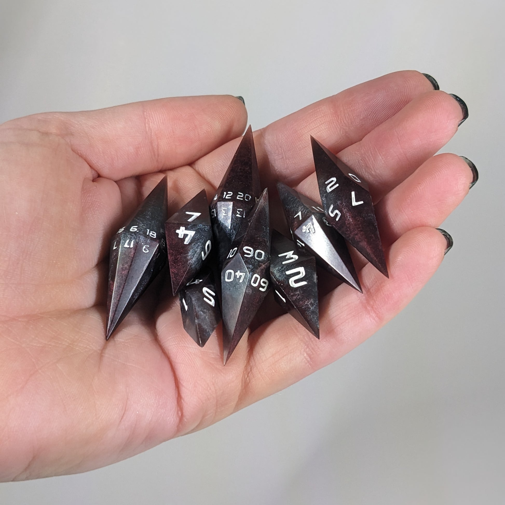 Close-up of deep maroon and dark silver with white numbers artisan dice resting in a palm on a seamless white backdrop. The handmade dice feature a distinct sharp diamond shape, with 2 d20s visible among the high-quality resin dice.