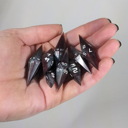 Close-up of deep maroon and dark silver with white numbers artisan dice resting in a palm on a seamless white backdrop. The handmade dice feature a distinct sharp diamond shape, with 2 d20s visible among the high-quality resin dice.
