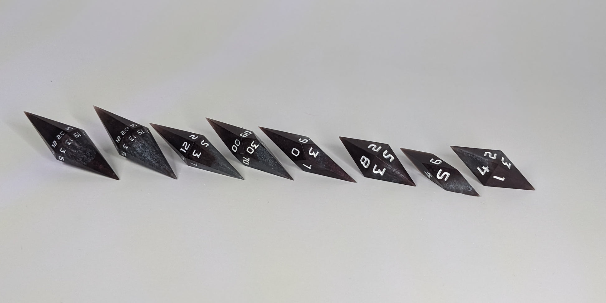 A side profile photograph of a complete set of deep maroon and dark silver with white numbers handmade dice arranged in a straight line. This eye-level view emphasizes the unique elongated and pointed diamond silhouette of the resin dice, showing the sharp geometry of the 2 d20s and matching artisan dice.