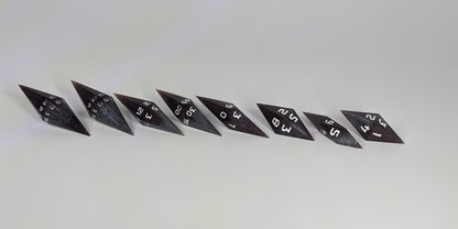 A side profile photograph of a complete set of deep maroon and dark silver with white numbers handmade dice arranged in a straight line. This eye-level view emphasizes the unique elongated and pointed diamond silhouette of the resin dice, showing the sharp geometry of the 2 d20s and matching artisan dice.
