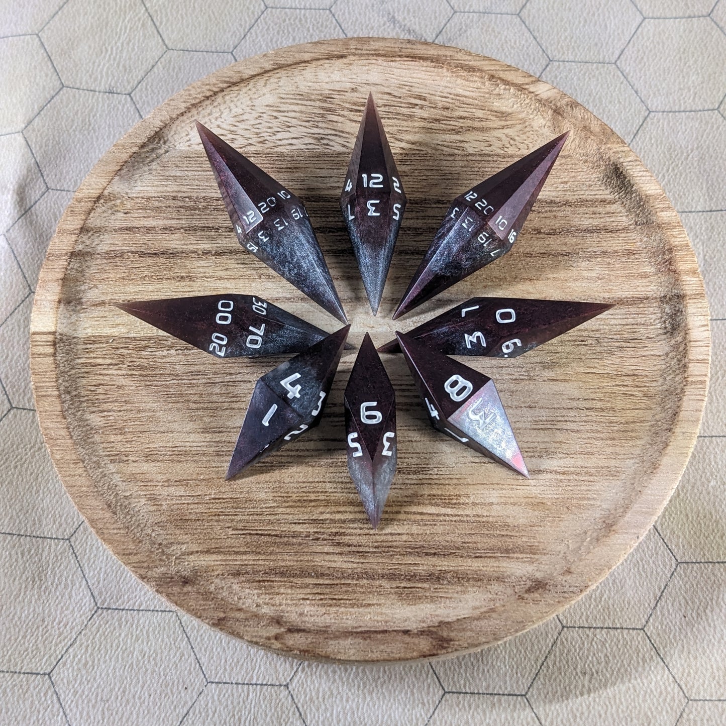 A set of deep maroon and dark silver with white numbers handmade dice arranged in a wooden circle organizer. The resin dice feature a unique pointed diamond shape with the tips facing outward, highlighting the sharp edges of the two d20 and other artisan dice in the collection. 
