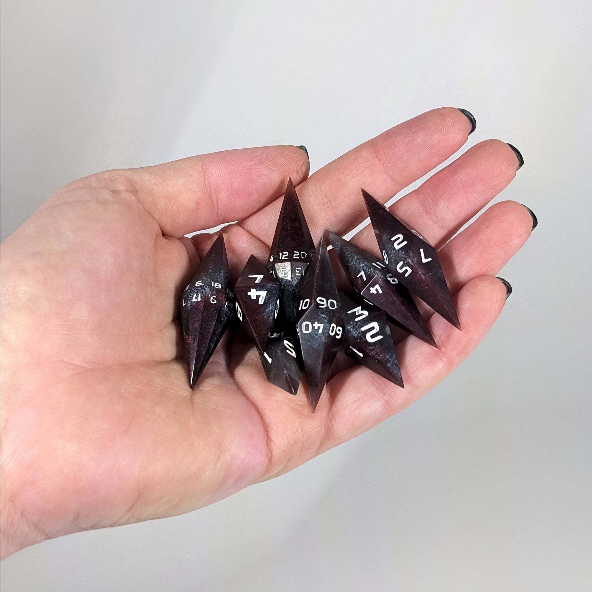 An 8 dice set of diamond style dice that are deep maroon and dark silver with white numbers are held in a hand. The image shows that the set fits easily in a hand, making them overall smaller, though sharper, than a standard set of dnd dice.