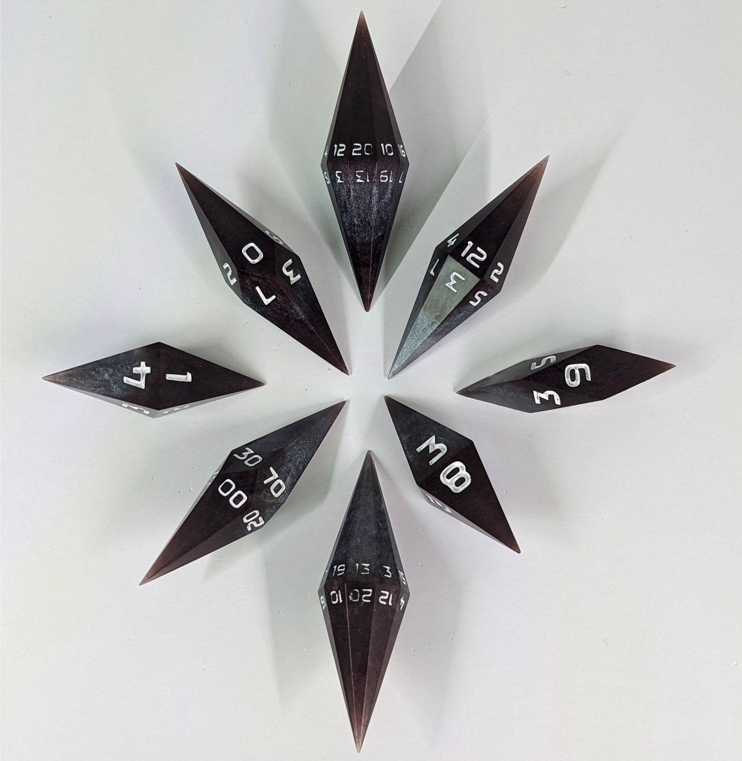 A complete set of diamond shaped resin dice featuring 2 d20s. These handmade dice are deep maroon and dark silver with white numbers and are clustered in a circle with the bottom points angled toward a central point on a white background.