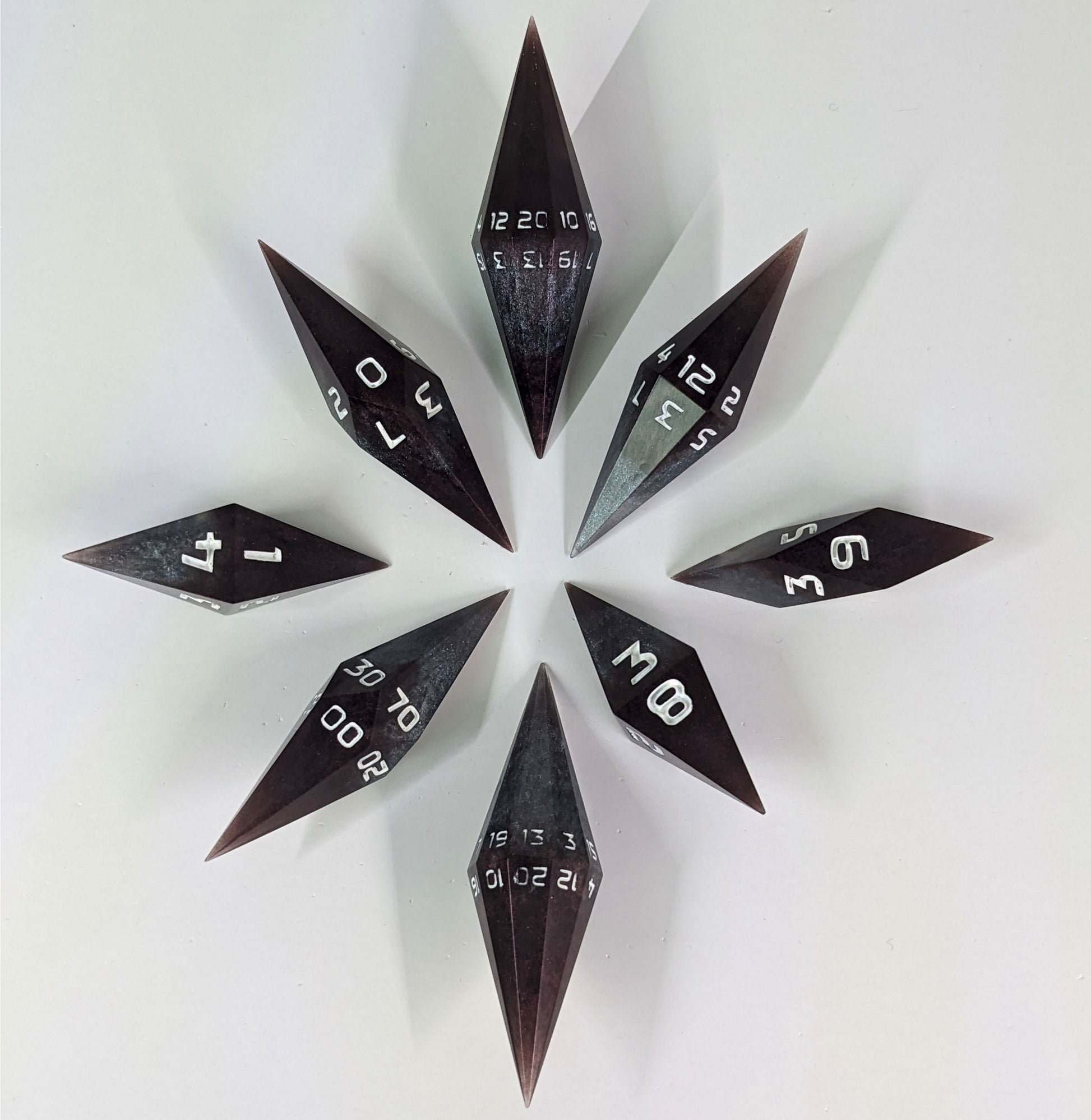 A complete set of diamond shaped resin dice featuring 2 d20s. These handmade dice are deep maroon and dark silver with white numbers and are clustered in a circle with the bottom points angled toward a central point on a white background.