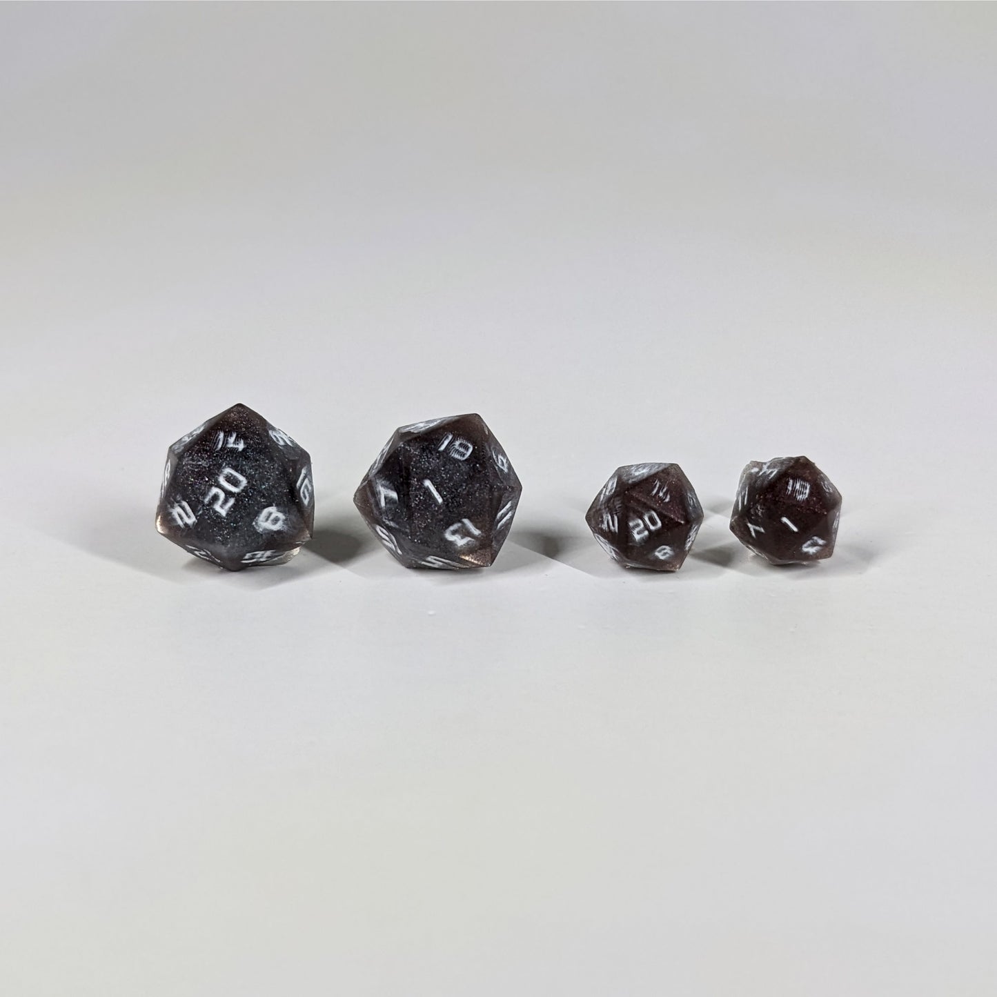 Two pairs of handmade d20 earrings with sterling silver hardware that are maroon and dark silver with white numbers on a white background sit in a line with the larger pair on the left and the smaller pair on the right. The earrings on the left of each pair centers the 20 face and the earrings on the right of each pair centers the 1 face.