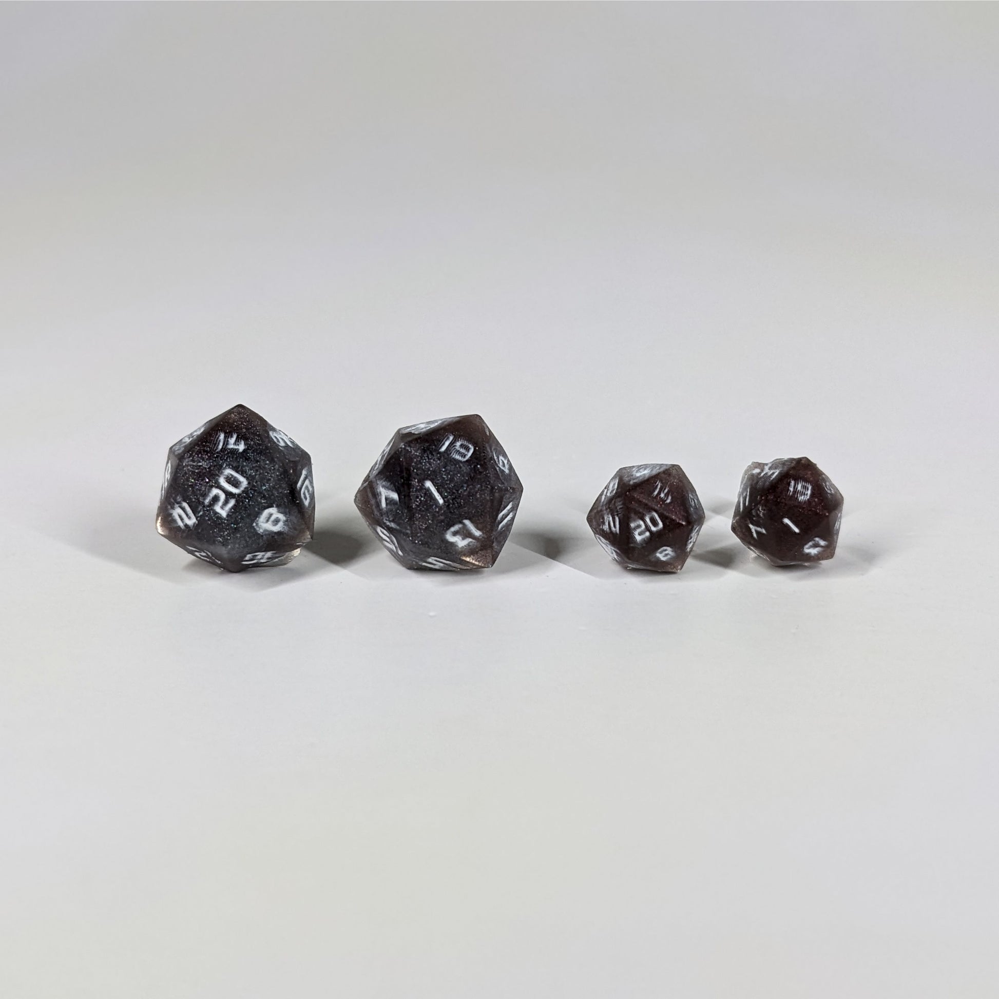 Two pairs of handmade d20 earrings with sterling silver hardware that are maroon and dark silver with white numbers on a white background sit in a line with the larger pair on the left and the smaller pair on the right. The earrings on the left of each pair centers the 20 face and the earrings on the right of each pair centers the 1 face.