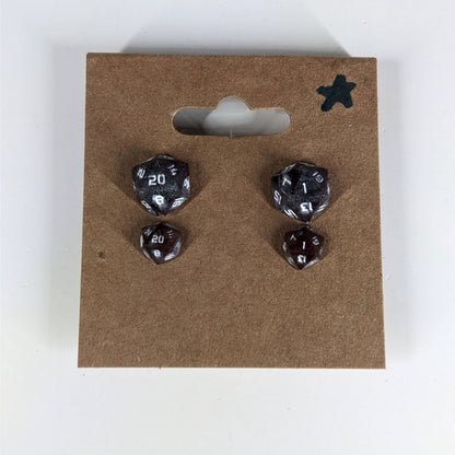 Two pairs of handmade d20 earrings with sterling silver hardware that are maroon and dark silver with white numbers on a kraft brown earring card with a star in the top right corner. The earrings on the left of each pair centers the 20 face and the earrings on the right of each pair centers the 1 face.
