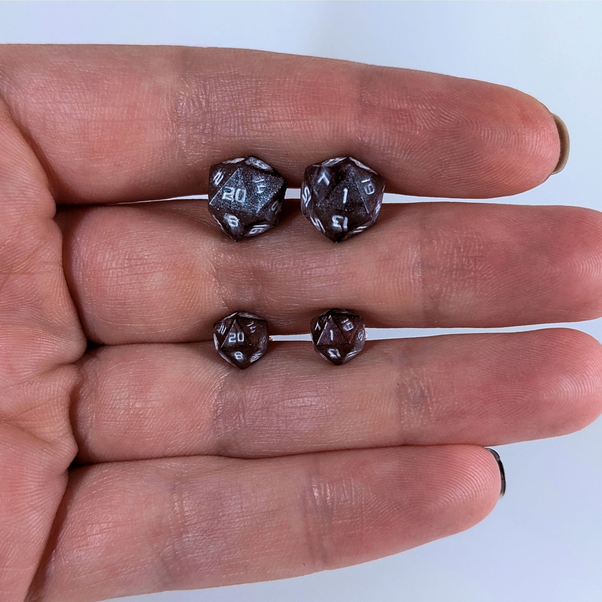 Two pairs of handmade d20 earrings with sterling silver hardware that are maroon and dark silver with white numbers held between the fingers of a flat hand. The larger pair is between the pointer and middle finger, the smaller between the middle and ring finger. The earrings on the left of each pair centers the 20 face and the earrings on the right of each pair centers the 1 face.