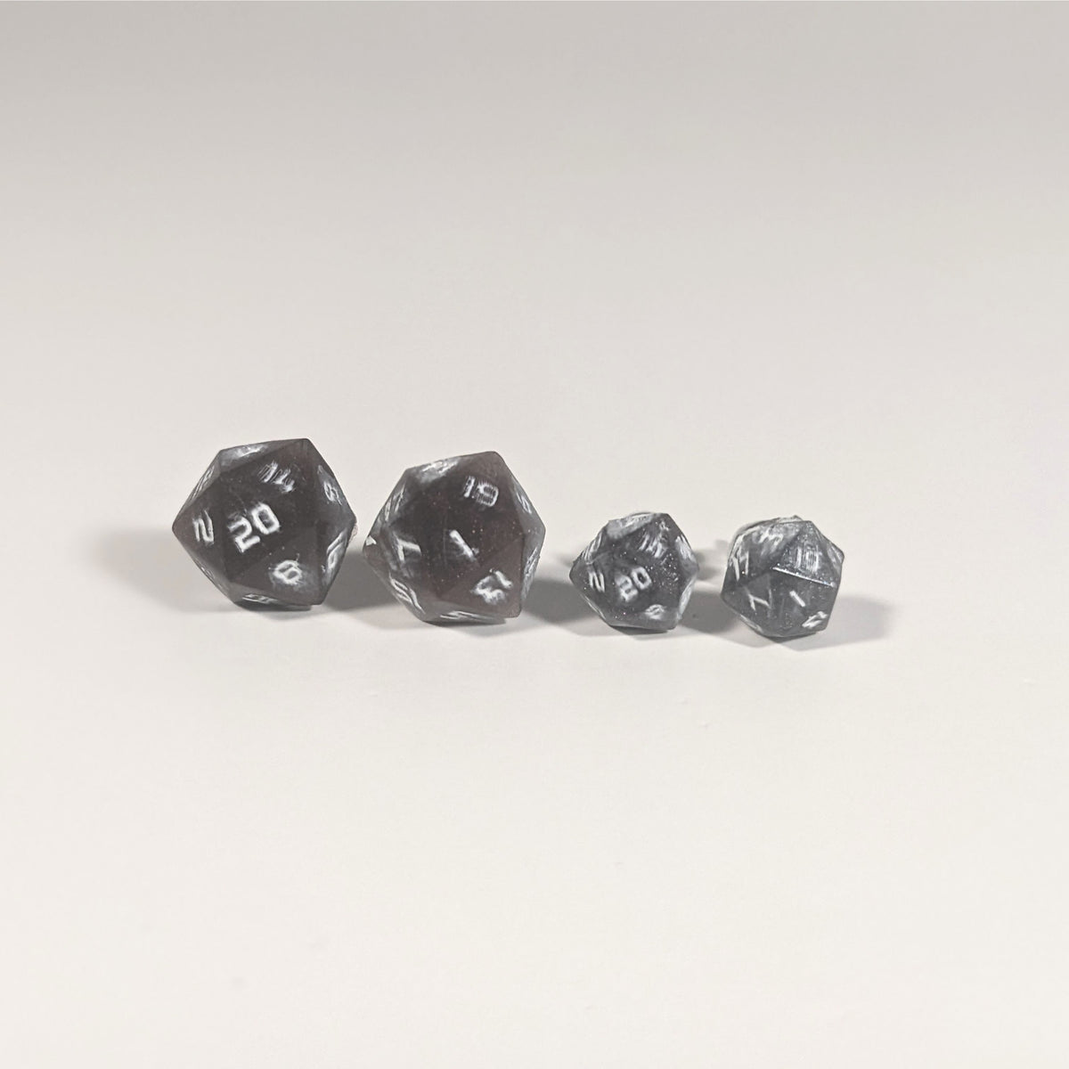 Two pairs of handmade d20 earrings with titanium hardware that are maroon and dark silver with white numbers on a white background sit in a line with the larger pair on the left and the smaller pair on the right. The earrings on the left of each pair centers the 20 face and the earrings on the right of each pair centers the 1 face.