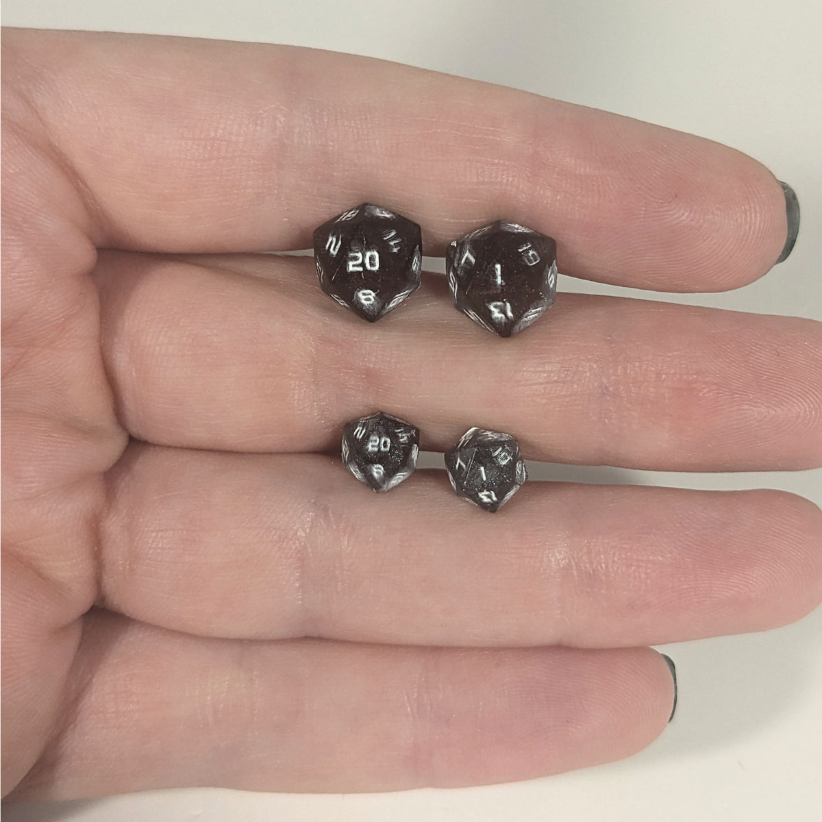 Two pairs of handmade d20 earrings with titanium hardware that are maroon and dark silver with white numbers held between the fingers of a flat hand. The larger pair is between the pointer and middle finger, the smaller between the middle and ring finger. The earrings on the left of each pair centers the 20 face and the earrings on the right of each pair centers the 1 face.