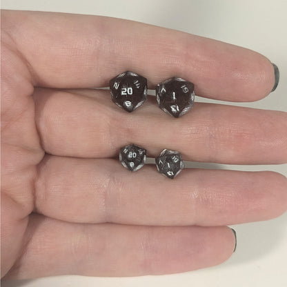 Two pairs of handmade d20 earrings with titanium hardware that are maroon and dark silver with white numbers held between the fingers of a flat hand. The larger pair is between the pointer and middle finger, the smaller between the middle and ring finger. The earrings on the left of each pair centers the 20 face and the earrings on the right of each pair centers the 1 face.