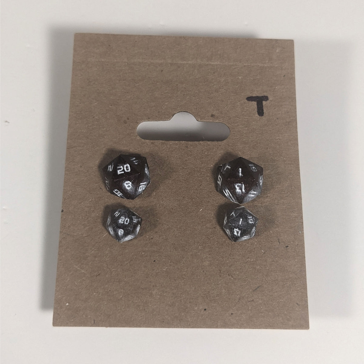 Two pairs of handmade d20 earrings with titanium hardware that are maroon and dark silver with white numbers on a kraft brown earring card with a T in the top right corner. The earrings on the left of each pair centers the 20 face and the earrings on the right of each pair centers the 1 face.
