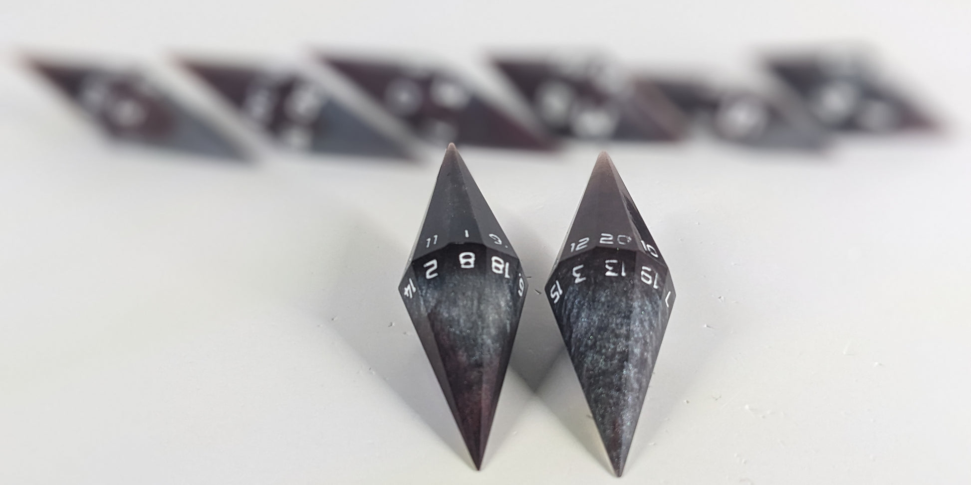 A stylized macro shot focusing heavily on 2 d20s from a deep maroon and dark silver with white numbers handmade dice set in the foreground. The sharp diamond edges of these front resin dice are crisp, while the rest of the matching artisan dice collection forms a softly blurred background, creating depth.