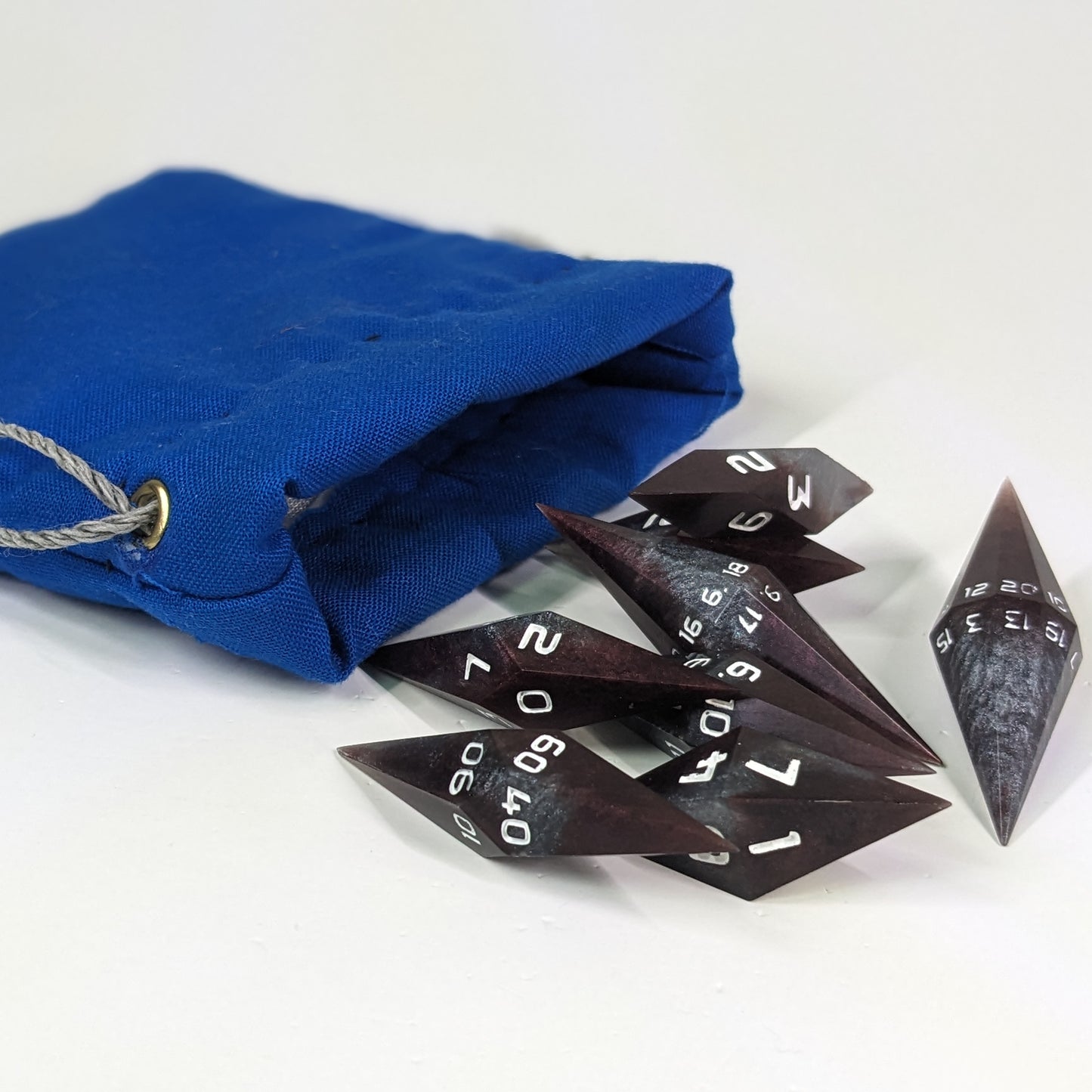Deep maroon and dark silver with white numbers diamond handmade dice spilling out of a blue drawstring bag on a white background. The set features 2 d20s and matching resin dice, showcasing the unique shape of these artisan dice.
