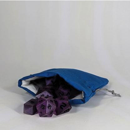 A small, handmade dice bag that is blue lays open with a set of purple dnd dice with black numbers spilling out of it on a white background.