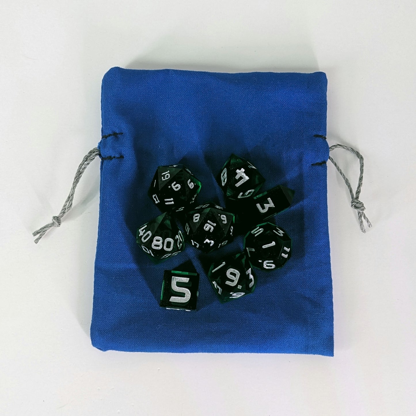 A small, handmade dice bag that is blue lays flat on a white background with a set of green dnd dice with silver numbers sitting on top of it.