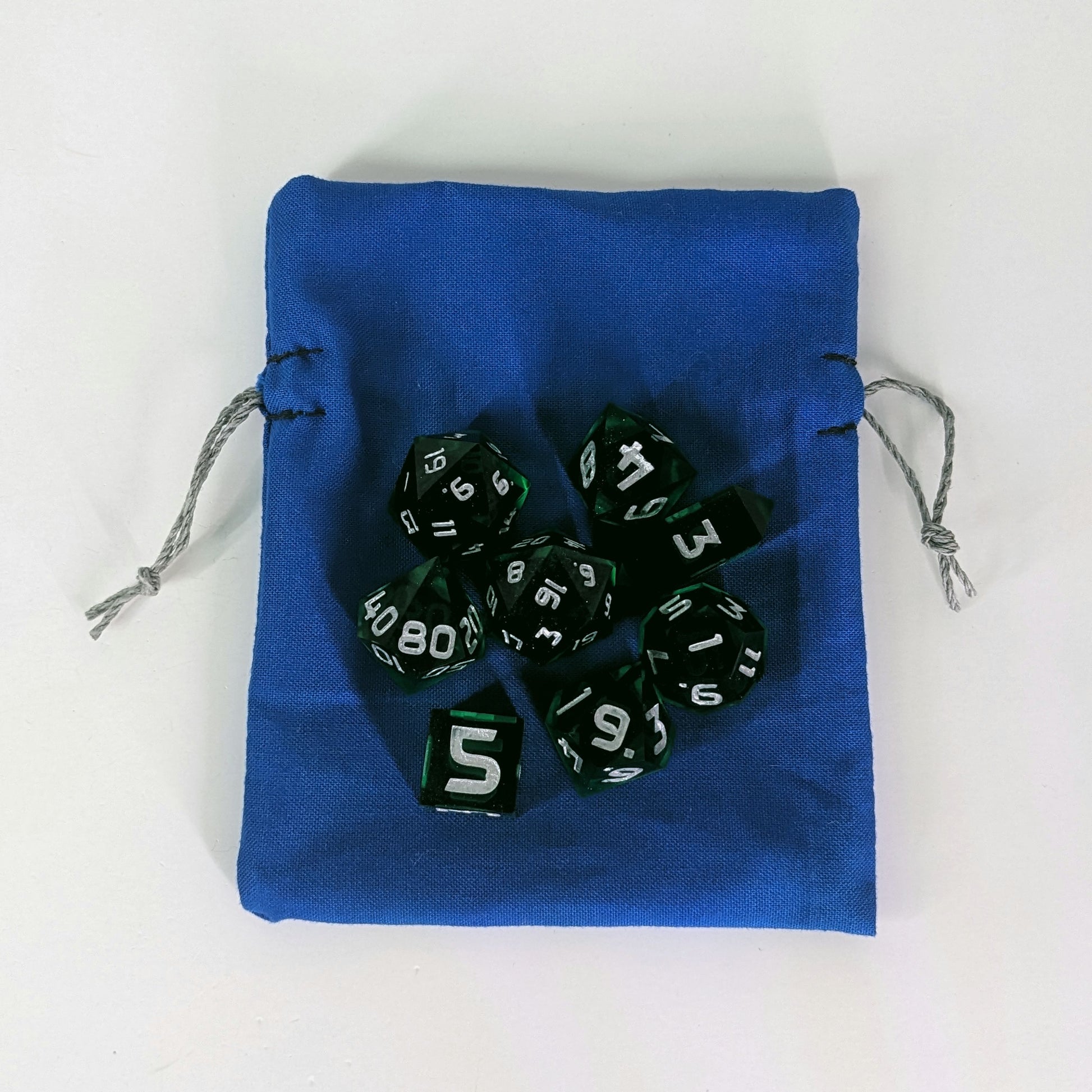 A small, handmade dice bag that is blue lays flat on a white background with a set of green dnd dice with silver numbers sitting on top of it.