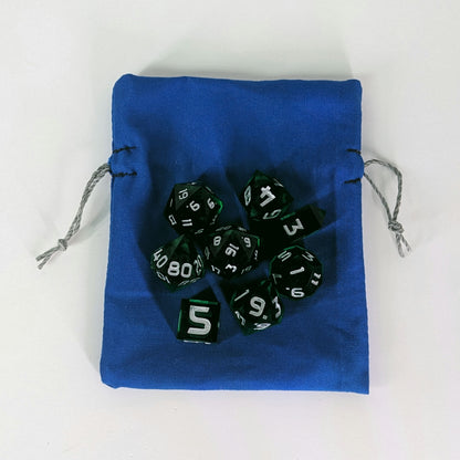 A small, handmade dice bag that is blue lays flat on a white background with a set of green dnd dice with silver numbers sitting on top of it.