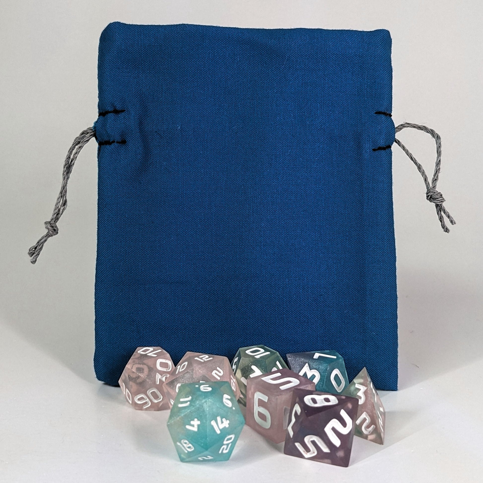 A small, handmade dice bag that is blue stands upright on a white background with a set of colorful dnd dice with white numbers sitting in front of it.